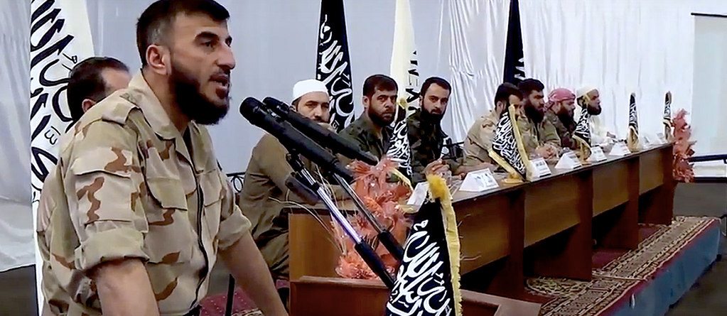 Zahran Alloush speaking at the declaration of the Unified Military Command in August 2014. Seated, left to right: Khaled Tafour, Abu Hamza al-Ghoutani, Yasser al-Qadri, Abdel-Nasr Shmeir, Abu Suleiman al-Zabadani, Samir Kaakeh, Said Darwish. Source: Rebel video.